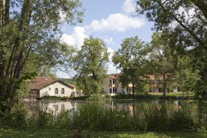 A serene lakeside setting with two buildings, one with a stone facade and the other with a white facade and red roof, surrounded by lush greenery and trees. - Logis Hotel la Source Bleue (Gudmont)