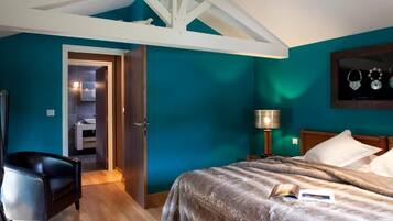 A bedroom with teal walls, a wooden ceiling, a bed with a fluffy blanket, a black chair, a bedside table with a lamp, and a framed jewelry display.