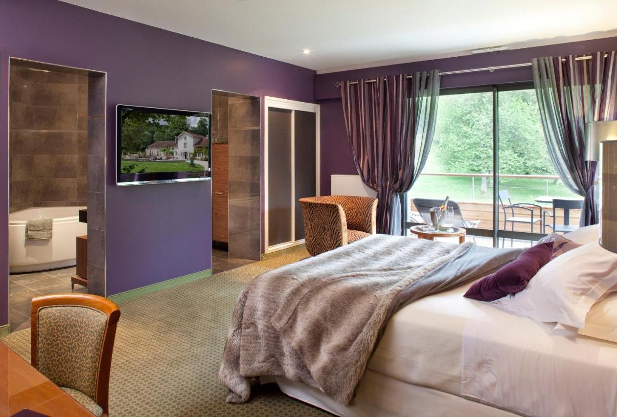 A hotel room with a large bed, a TV mounted on the wall, a seating area with a chair and table, and a view of the outdoors through the window.