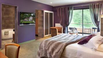 A hotel room with a large bed, a TV mounted on the wall, a seating area with a chair and table, and a view of the outdoors through the window.