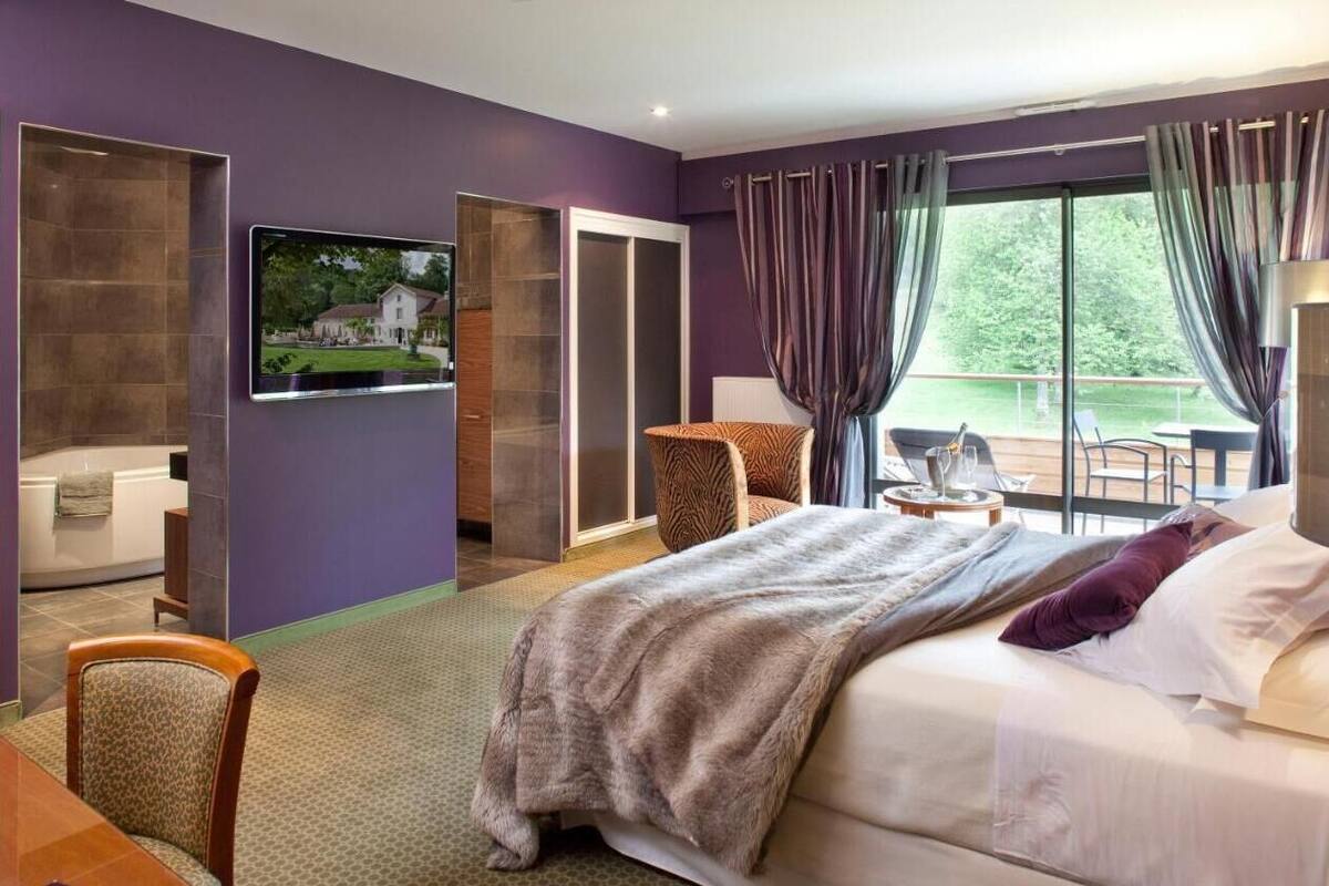 A hotel room with a large bed, a TV mounted on the wall, a seating area with a chair and table, and a view of the outdoors through the window.