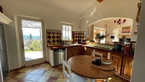 Une cuisine avec une table en bois, des chaises blanches, un lave-vaisselle et une vue sur l’extérieur, visible à travers la porte.