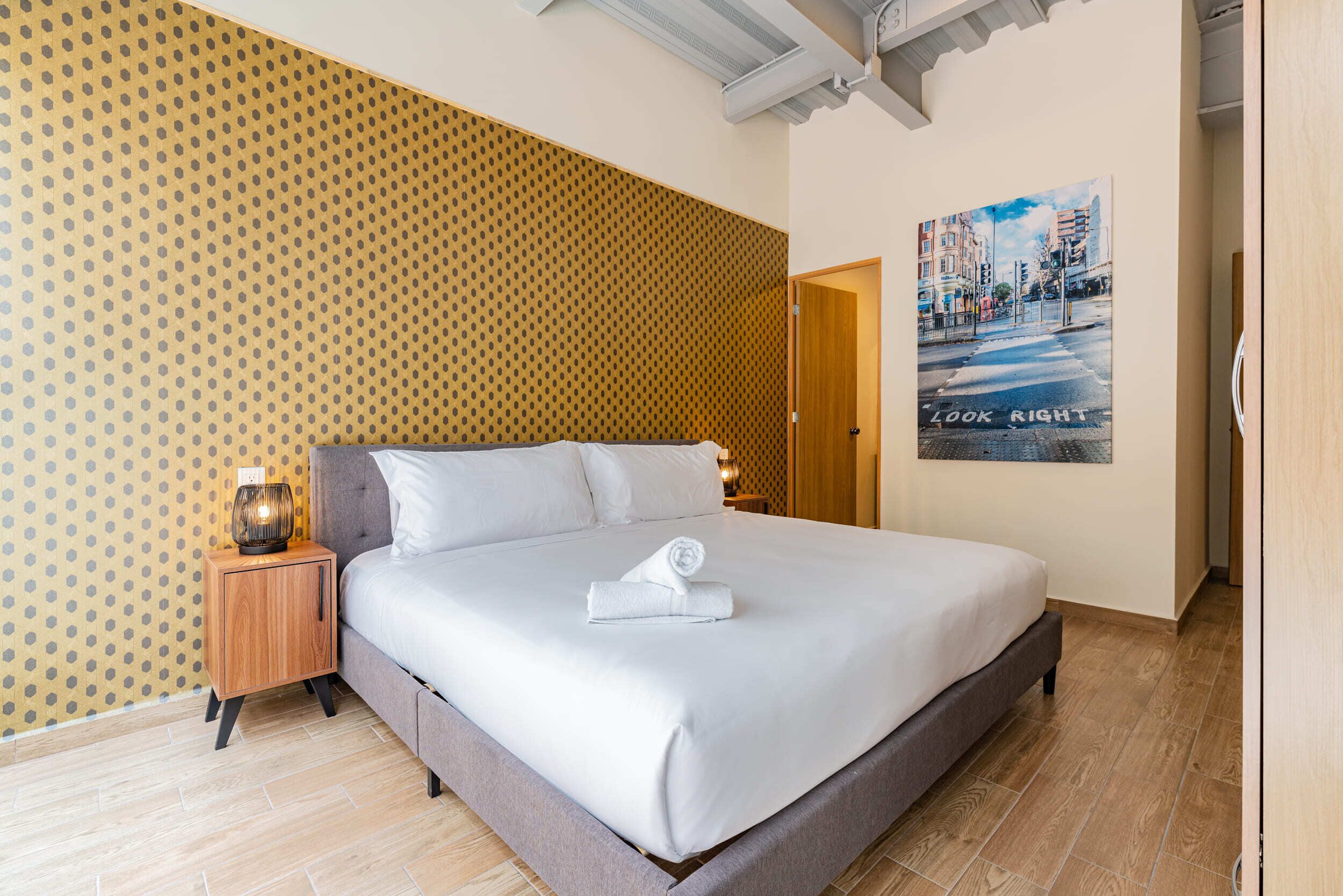 A neatly made bed with a rolled towel, a wooden nightstand with a lamp, a poster on the wall, and a patterned wallpaper.