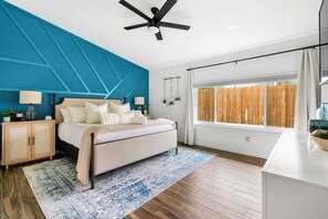 A modern bedroom with a bed, nightstands, a ceiling fan, a large window, and a view of a wooden fence.