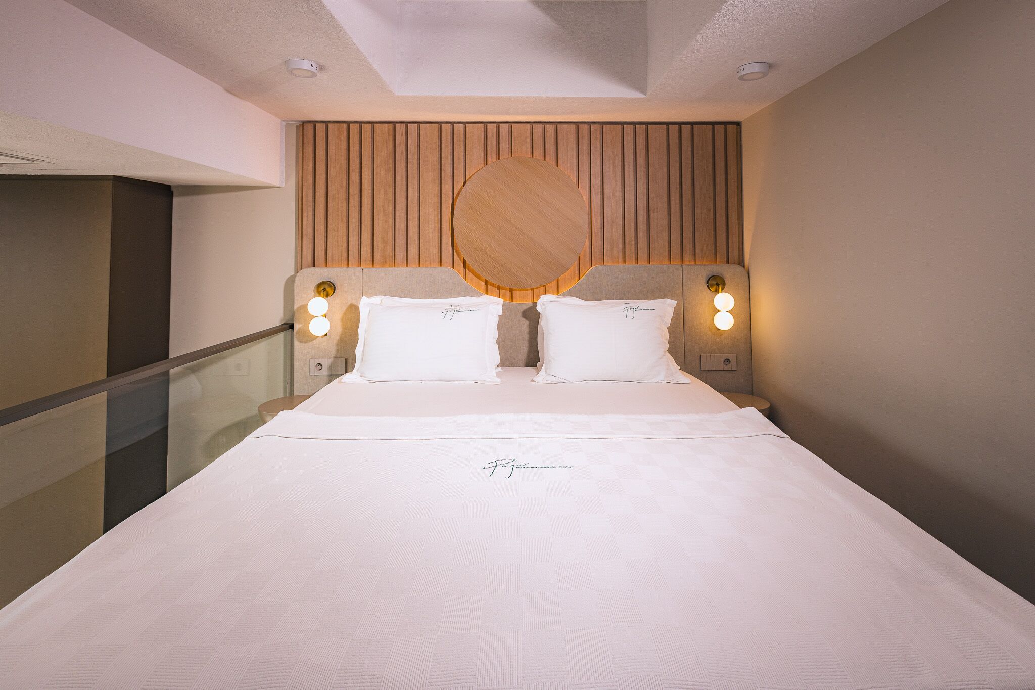 Quarto de hotel moderno com uma cama grande, duas luminárias de cabeceira e uma cabeceira de madeira.