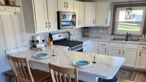 A kitchen with a large island, wooden chairs, a stainless steel trash can, and white cabinets. - Serenity Lakeside Cottage (Edinboro)