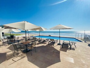 Zona junto a la piscina con sillones de descanso y sombrillas, con vista a un cielo azul despejado y al océano.