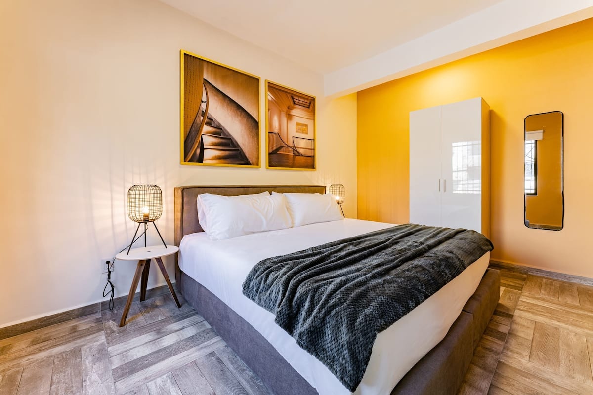 A modern hotel room with a large bed, a wooden floor, a wardrobe, and a wall-mounted mirror.