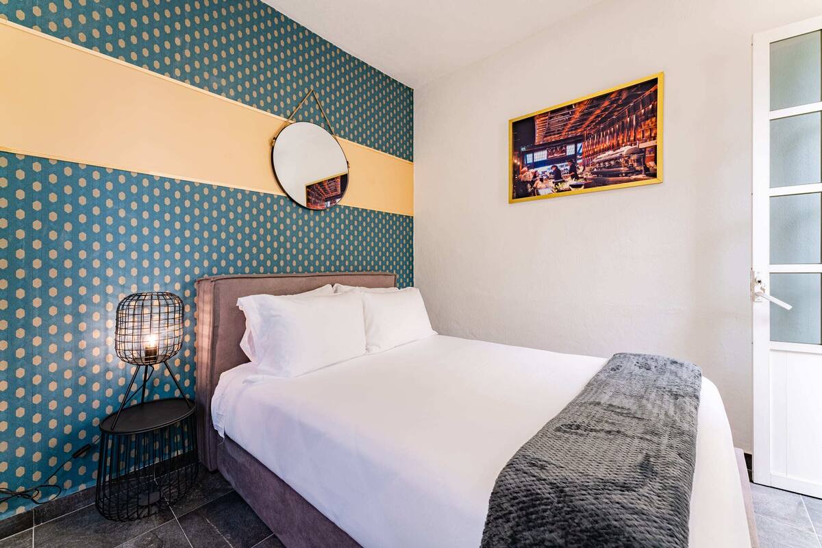 A neatly made bed with white linens, a round mirror on the wall, and a patterned wallpaper.