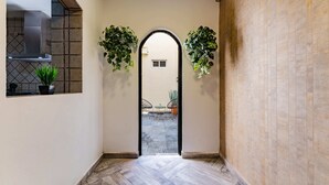 Un pasillo interior con suelo de baldosas, una puerta negra que da a un espacio exterior, y dos plantas en macetas a cada lado de la puerta.