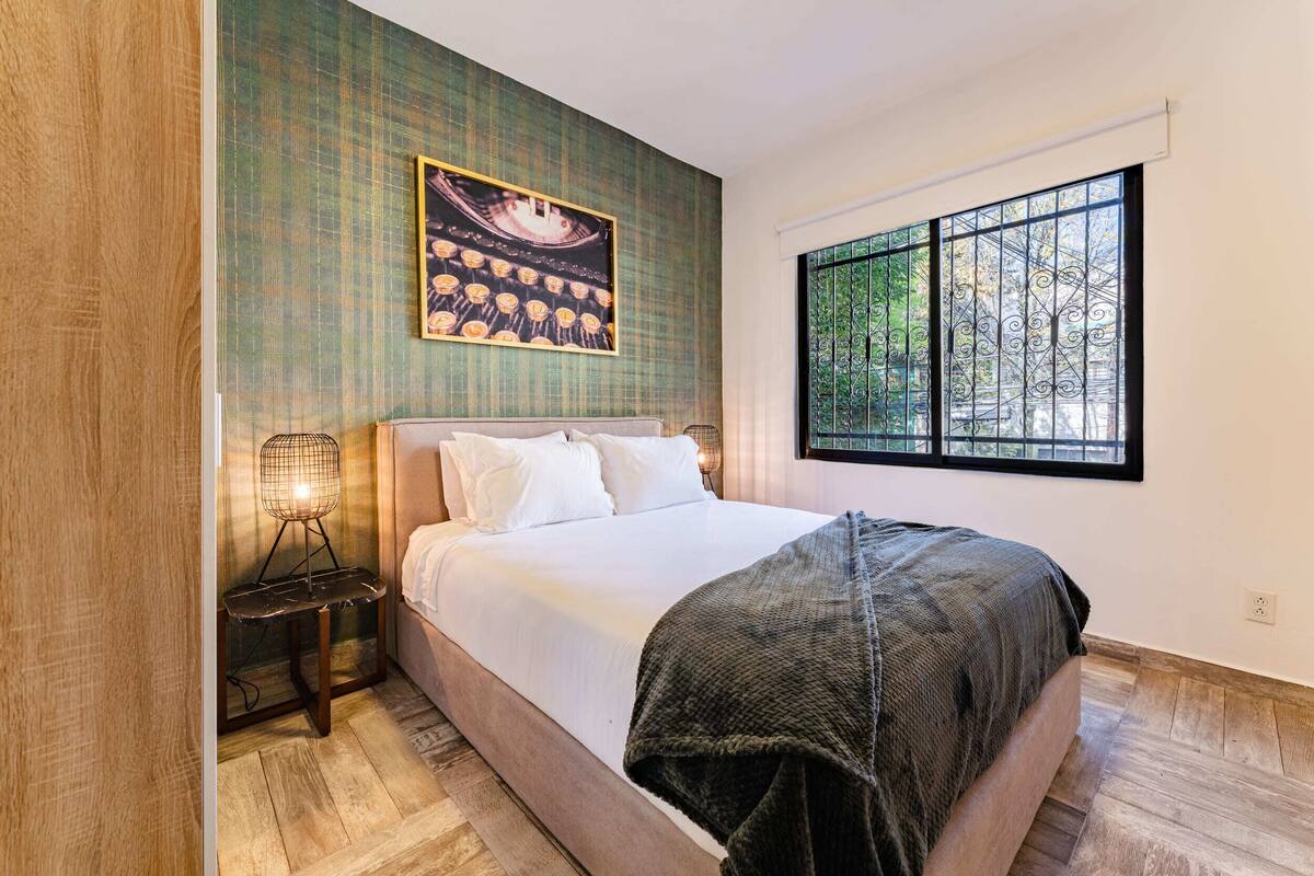 A bedroom with a large bed, a nightstand, a wall-mounted picture, and a window with a view of trees.