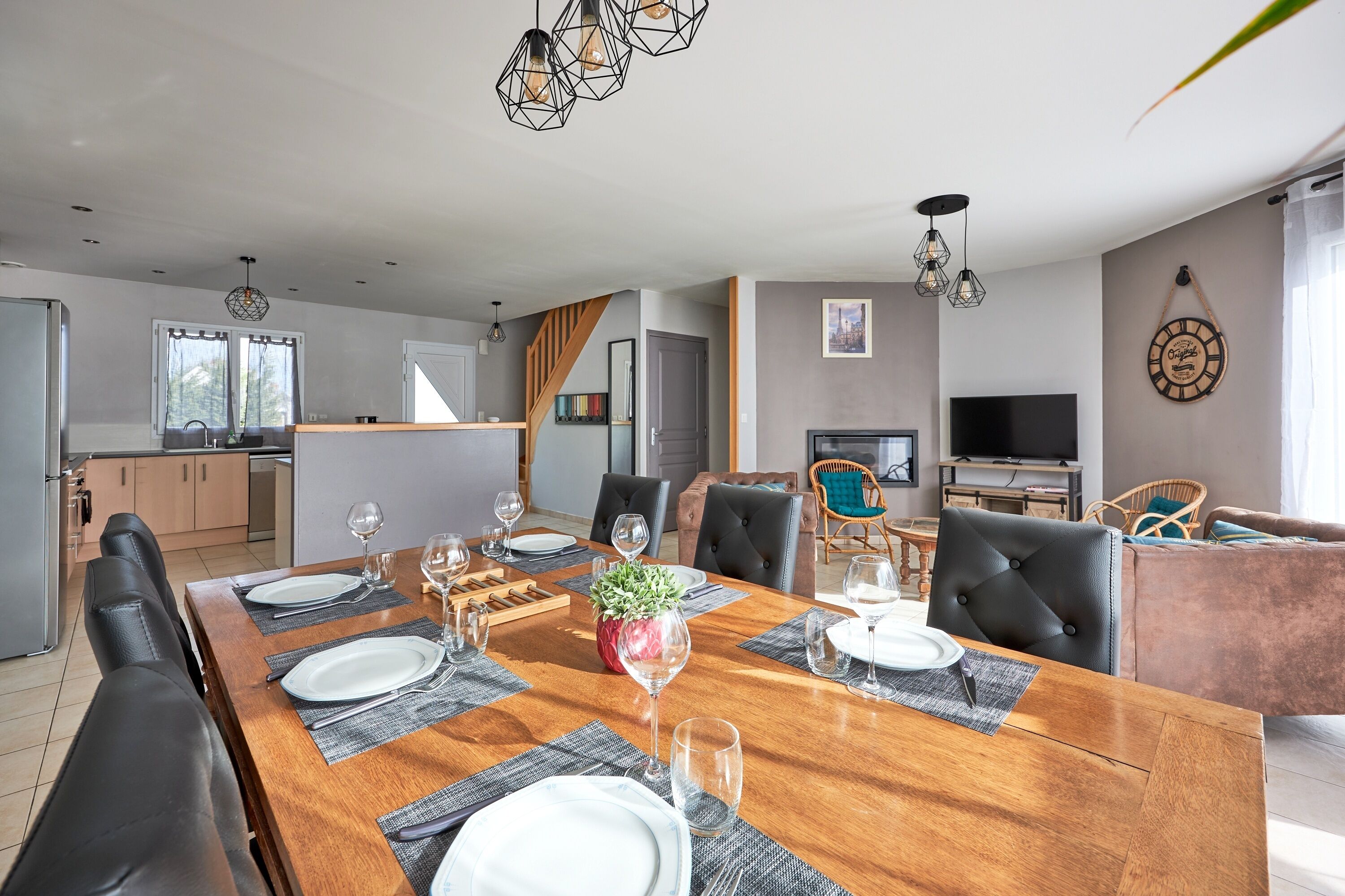 Un espace repas moderne avec une table en bois dressée pour six personnes, entourée de chaises en cuir. On aperçoit, au fond, une cuisine avec un plan de travail en bois et un coin repas équipé d’un téléviseur et de sièges confortables.