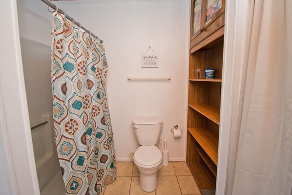 A bathroom with a patterned shower curtain, a toilet, a shelf with toiletries, and a sign on the wall. - Speight House, Soundfront House in Historical Beaufort, NC! (Beaufort)