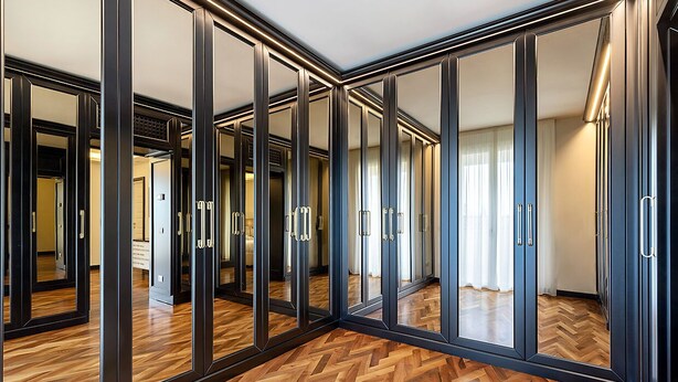 Quarto com portas de armário espelhadas, soalho em parquet de madeira e vista para outro quarto através dos espelhos.