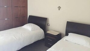 Habitación de hotel con dos camas individuales, un armario de madera, una mesita de noche pequeña y una lámpara de pared.