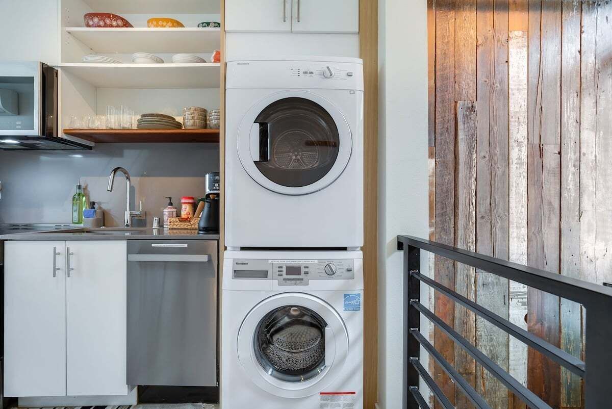 A compact kitchen with a dishwasher, a stacked washer and dryer, and wooden shelving.