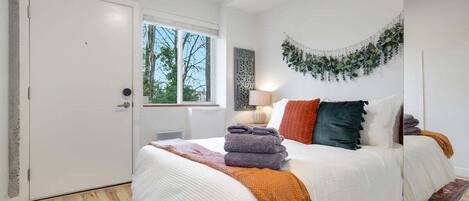 A bedroom with a large bed, a window with a view of trees, a door, and a wall-mounted plant decoration.
