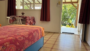 Ein Schlafzimmer mit Bett, Tisch und Stuhl. Ein Fenster mit Vorhängen, eine zur Außenseite führende Tür und Blick ins Grüne.
