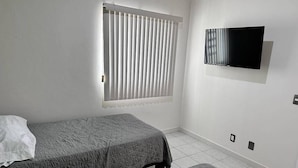 A bedroom with a bed, a window with blinds, a wall-mounted TV, and a tiled floor. - Condos PDS (Mazatlán)