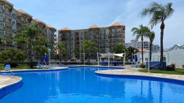 A swimming pool surrounded by palm trees and lounge chairs, with multi-story buildings in the background. - Condos PDS (Mazatlán)