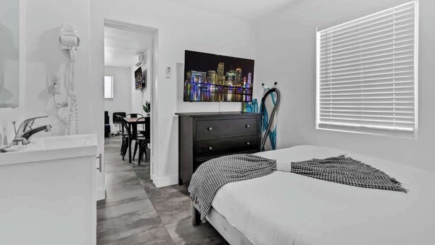 A bedroom with a bed, a dresser, a nightstand, a window with blinds, a painting of a cityscape, and a bathroom with a hairdryer.