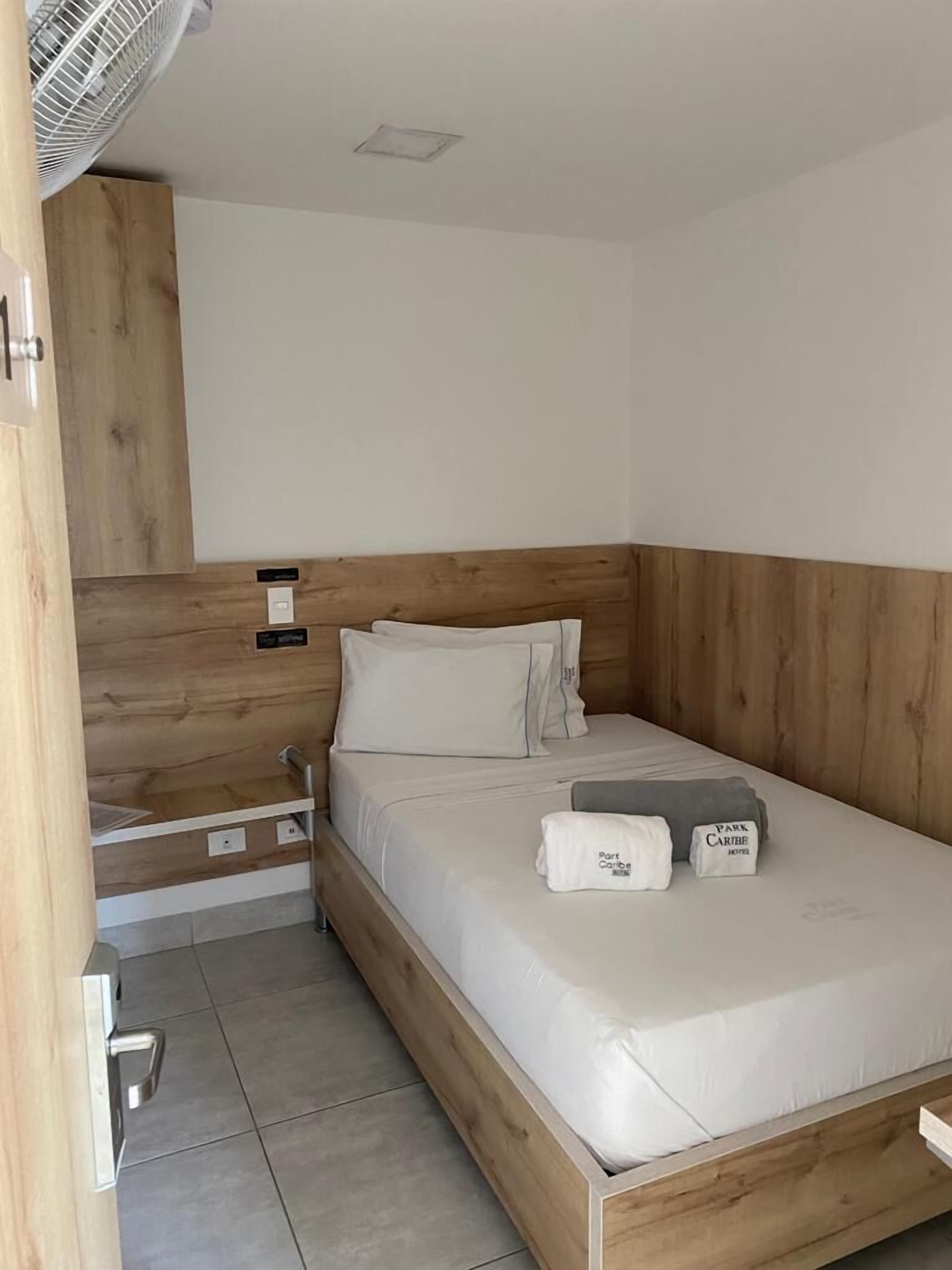 Una cama individual con cabecera de madera, una mesita de noche y una mesita auxiliar. Hay dos almohadas y una toalla doblada sobre la cama.