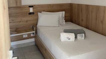 Una cama individual con cabecera de madera, una mesita de noche y una mesita auxiliar. Hay dos almohadas y una toalla doblada sobre la cama.