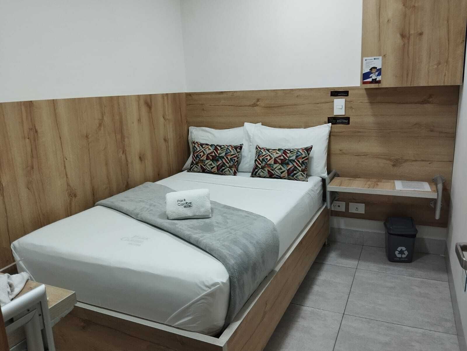 Un lit bien fait, avec une couverture grise et deux coussins à motifs, une table de chevet avec une lampe, une petite poubelle et une tête de lit en bois.
