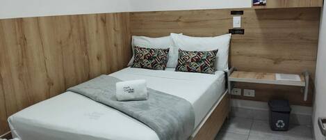 Un lit bien fait, avec une couverture grise et deux coussins à motifs, une table de chevet avec une lampe, une petite poubelle et une tête de lit en bois.
