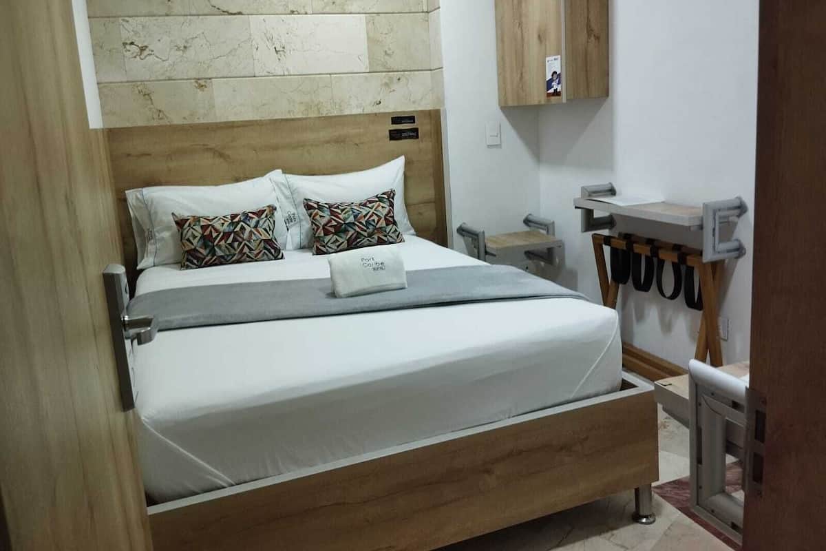Ein Schlafzimmer mit einem Bettgestell aus Holz, weißer Bettwäsche und gemusterten Kissen. Es gibt einen kleinen Nachttisch mit einer Lampe, einen Holzschrank über dem Bett und ein Metallgestell mit schwarzen Riemen.
