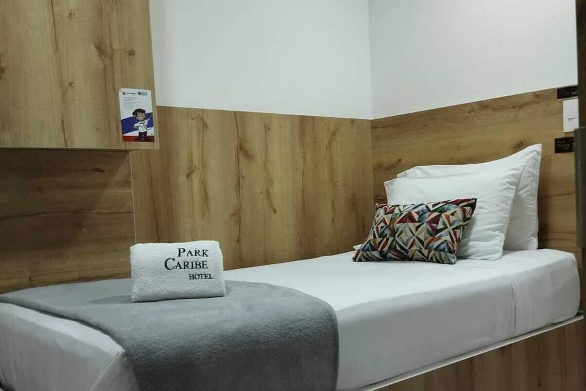 Ein Einzelbett mit grauer Decke, ein Kissen mit dem Aufdruck 'Park Caribe Hotel' und ein farbenfrohes geometrisches Kissen.
