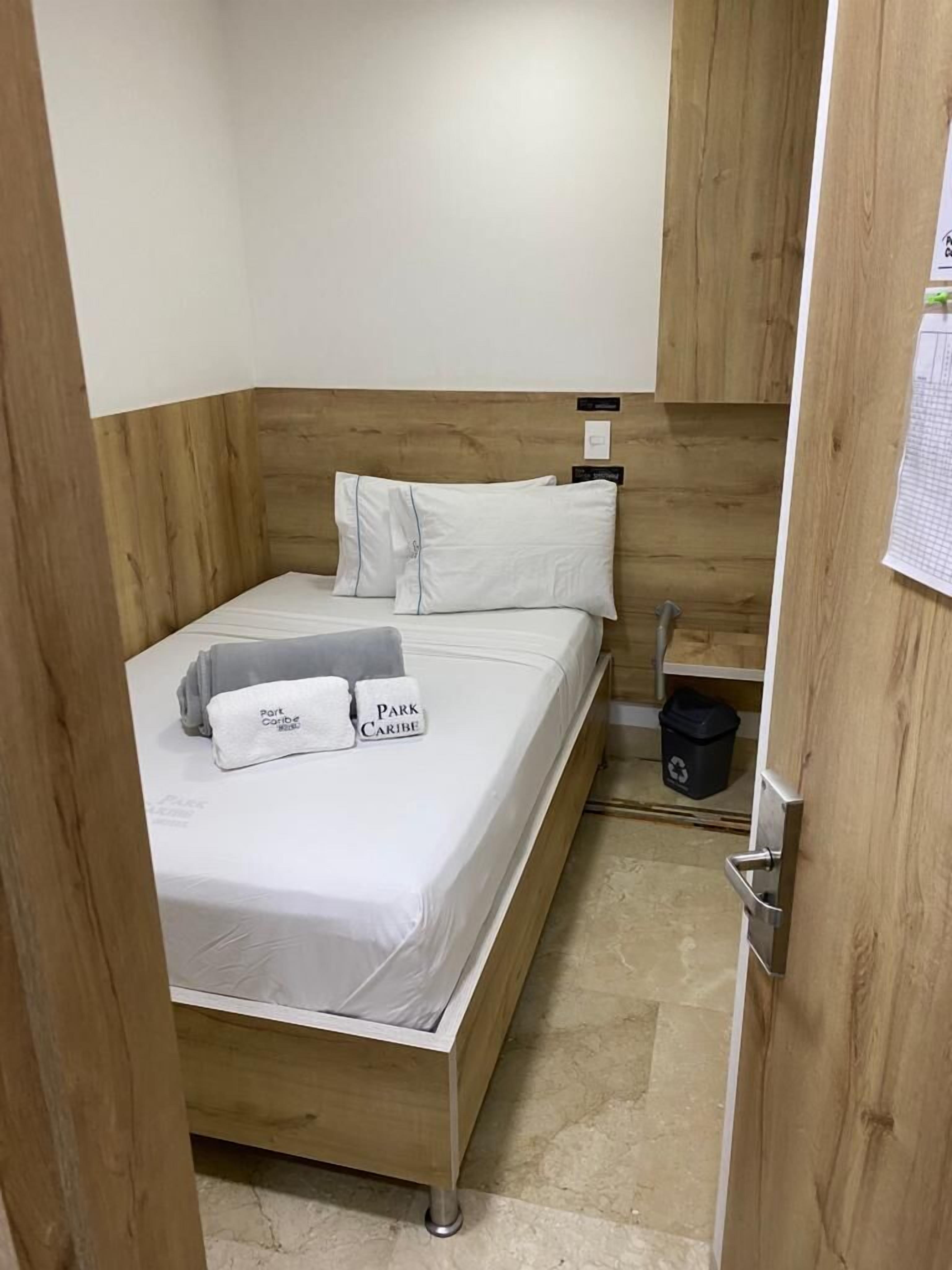 Una cama bien tendida con sábanas blancas y dos almohadas, un cabecero de madera, una mesita de noche pequeña y un cubo de basura.