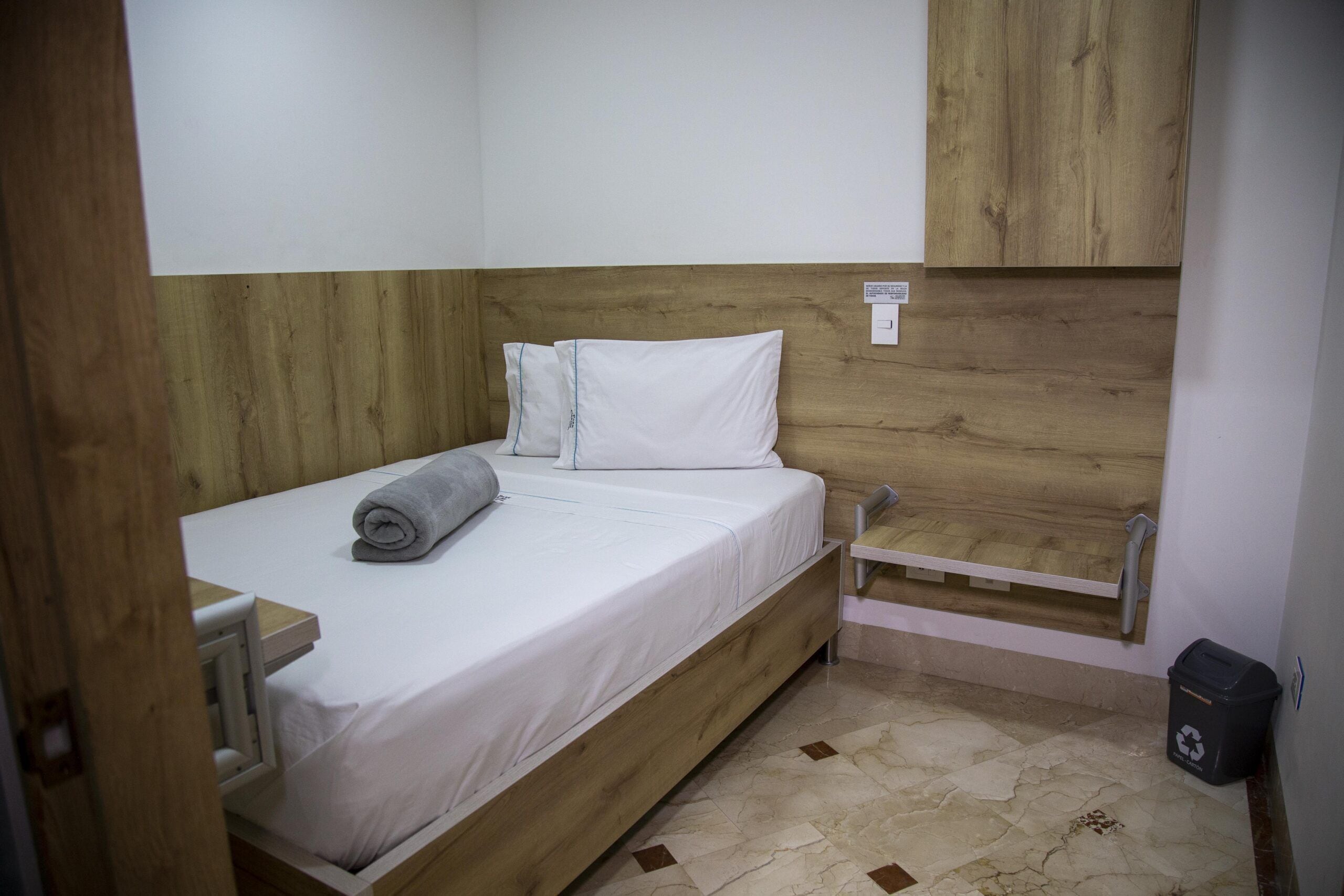 A small, single-bed room with a wooden headboard, a white mattress, and a grey towel.