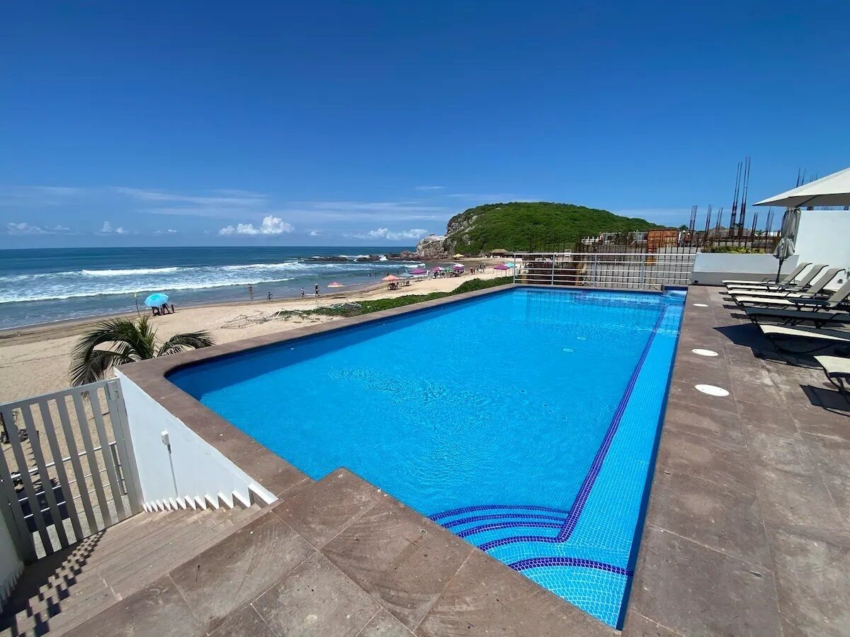 A rectangular swimming pool with a tiled edge, adjacent to a beach with a rocky cliff and a clear blue sky.
