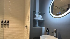 A modern bathroom with a glass shower enclosure, a round mirror, a white sink, and a wooden vanity. - Amae21 Lofts (San Pedro Garza García)