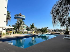 Una piscina con sillones de descanso, palmeras y un edificio con una torre circular.