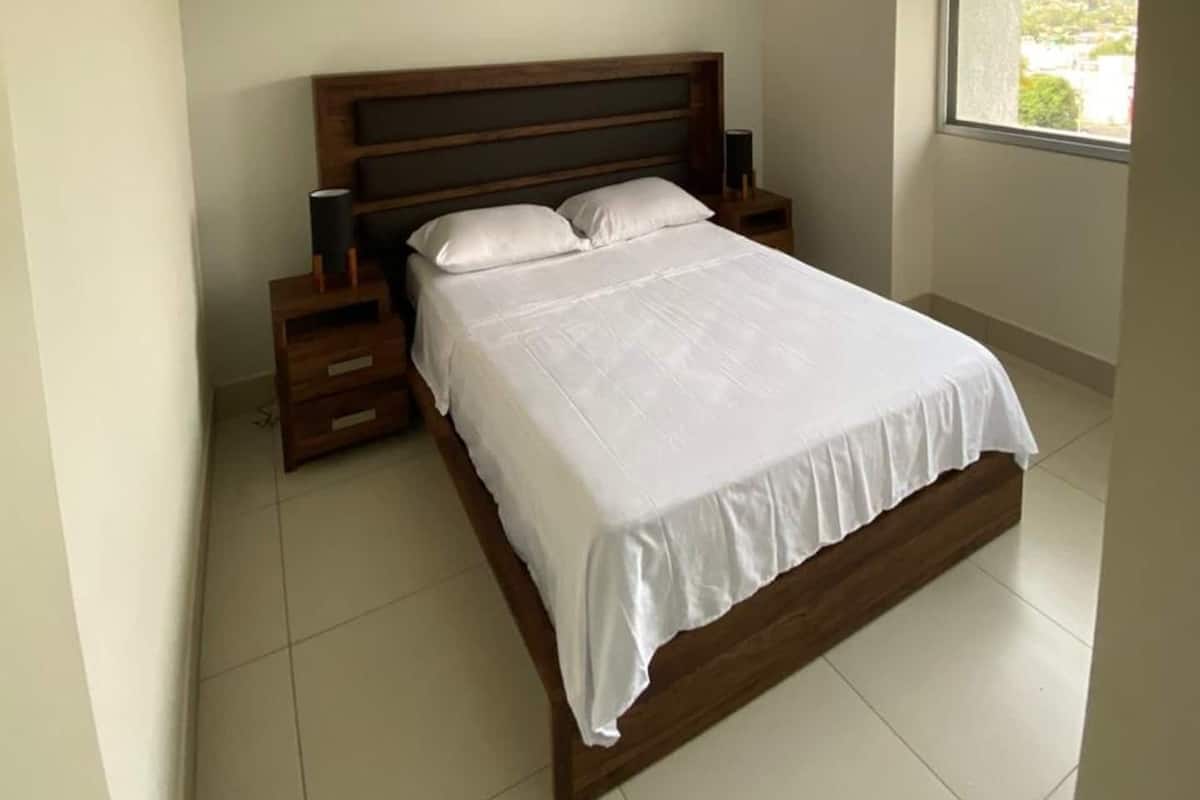 A bedroom with a wooden bed, white bedding, a nightstand, a bedside table, and a window with a view.