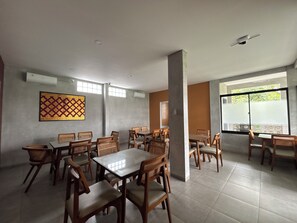 Uma área de refeições com várias mesas e cadeiras, aparelhos de ar condicionado e uma janela grande com vista.