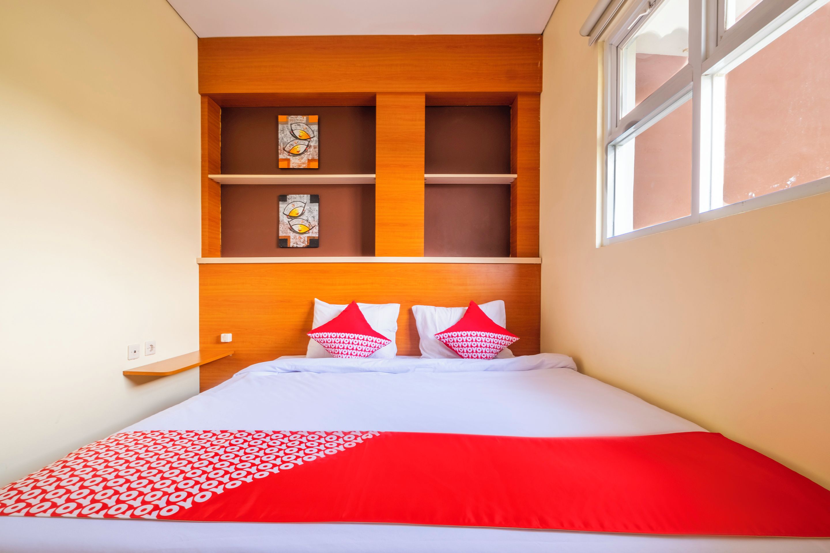 A hotel room with a bed, two pillows, a red and white blanket, a shelf with artwork, and a window.