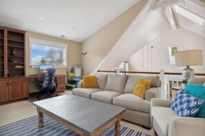 A spacious living room with a high ceiling, a large window, a sofa set, a dining table, and a wooden cabinet.