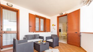 Un balcón con mobiliario de mimbre, suelo de baldosas y puertas de madera.