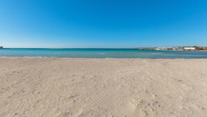 Une plage de sable fin bordée d’une eau d’un bleu limpide, avec une côte lointaine ponctuée de bâtiments.