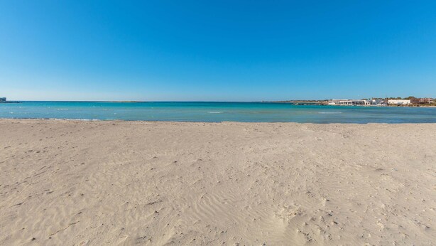 Una playa arenosa con agua azul cristalina y una costa lejana con edificios.