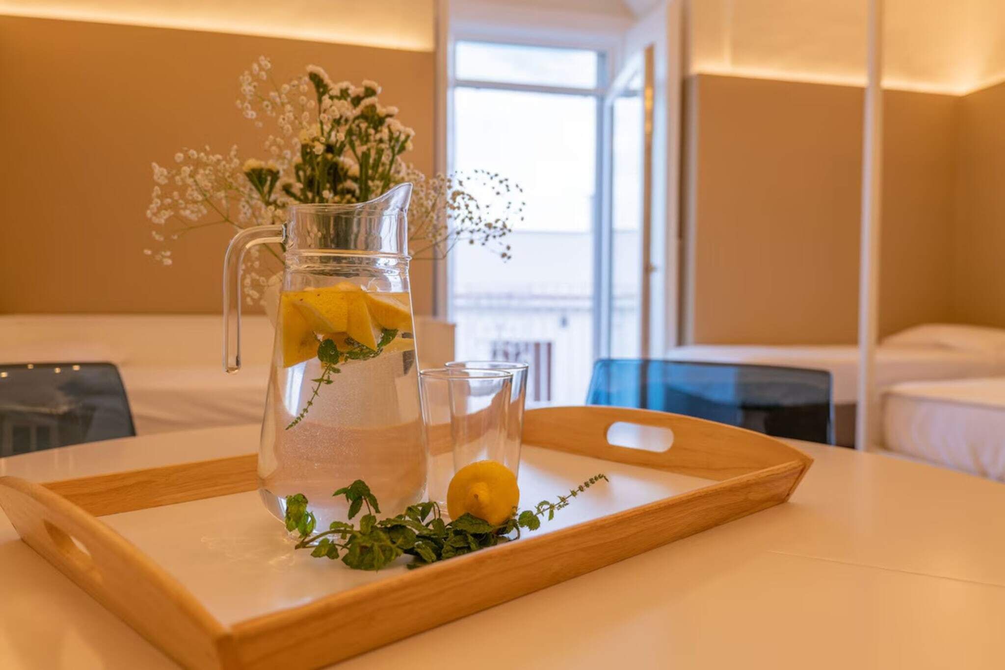 A clear glass pitcher with a lemon slice and herbs on a wooden tray, accompanied by two glasses, in a hotel room with a bed and a window in the background.