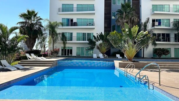 Une piscine entourée de chaises longues et de palmiers, le tout bordé de bâtiments modernes dotés de balcons.
