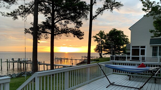 A wooden deck with a hammock, overlooking a sunset over the water with a dock in the background.