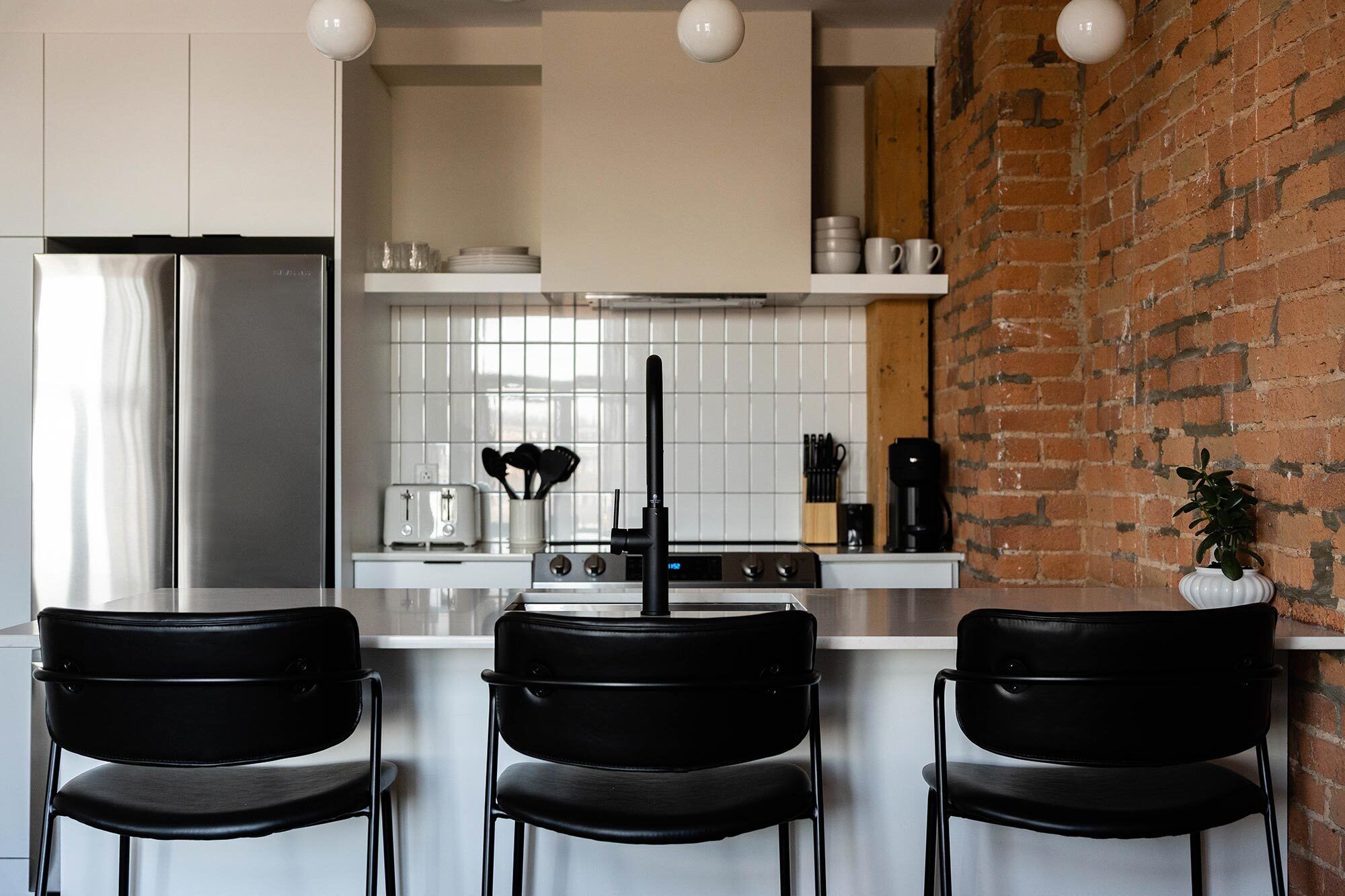 Een moderne keuken met een roestvrijstalen koelkast, witte kastjes, een bakstenen muur en zwarte stoelen.