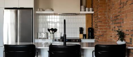 Een moderne keuken met een roestvrijstalen koelkast, witte kastjes, een bakstenen muur en zwarte stoelen.