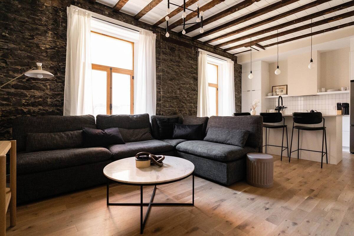 A modern living room with a dark grey sectional sofa, a round coffee table, and a kitchen area with a bar stool and a trash can.