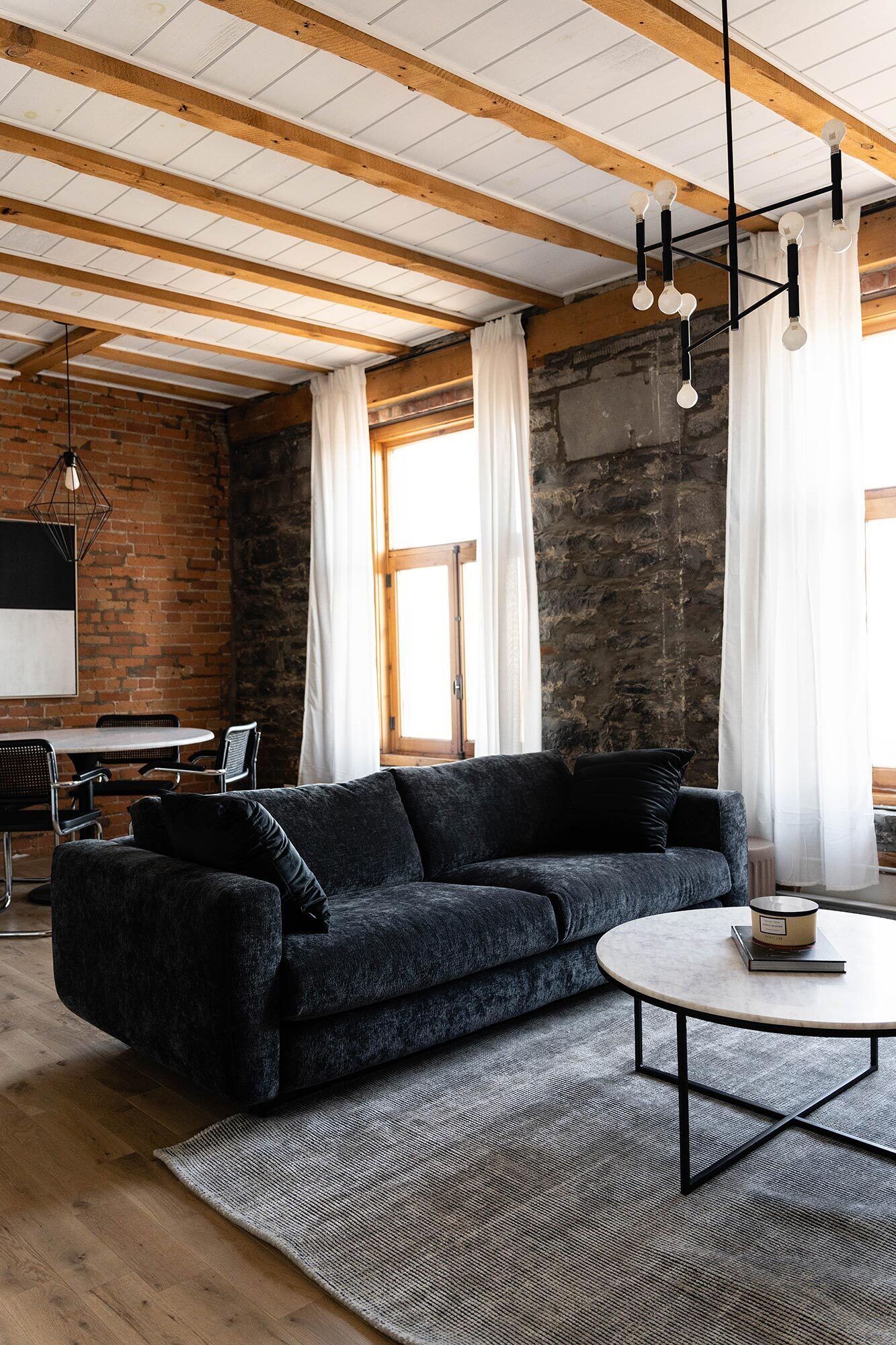 Ein modernes Wohnzimmer mit einem dunkelgrauen Ecksofa, einem runden Couchtisch und einem TV-Schrank aus schwarzem Metall. Im Raum sind Holzbalken sichtbar, eine Steinwand und ein großes Fenster mit weißen Vorhängen.